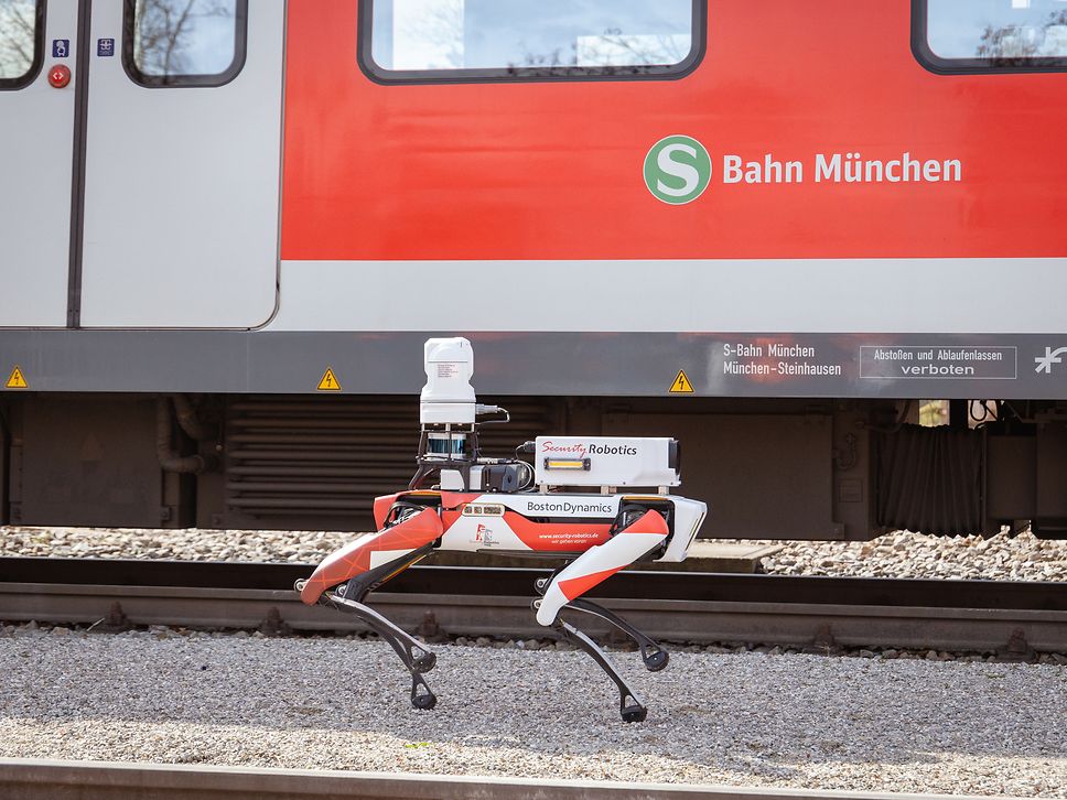 Roboterhund läuft vor Münchener S-Bahn am Gleis entlang